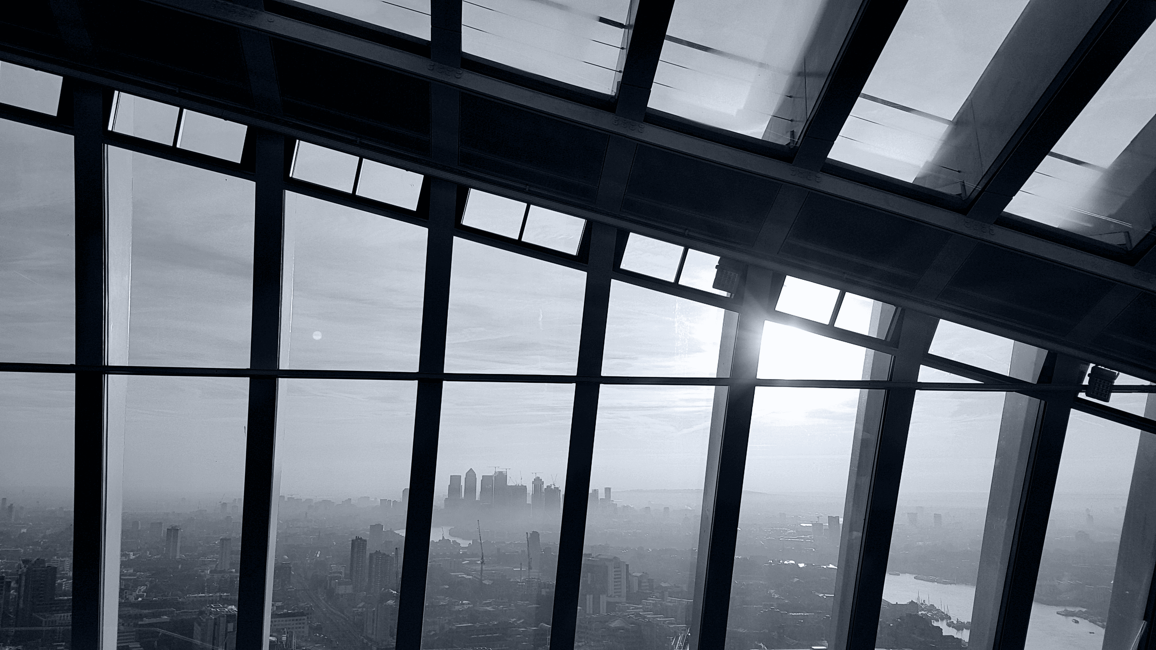 London skyline seen through the diagonal steel and glass roof of the Sky Garden, with sunlight falling through the windows